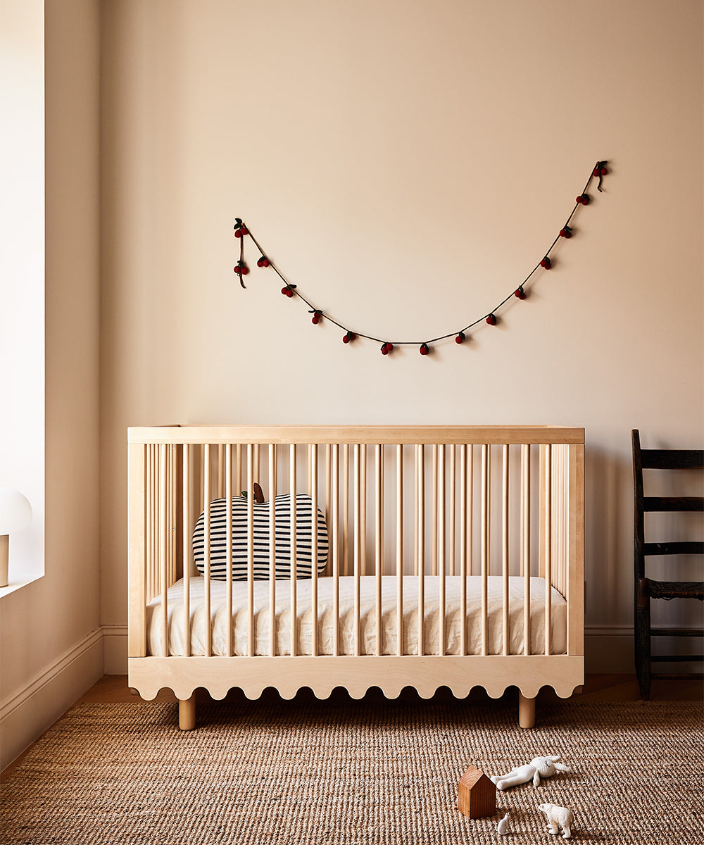 Minimalist nursery with a wooden crib, Apple Pillow, wall garland, black chair, wooden toys, and woven rug.