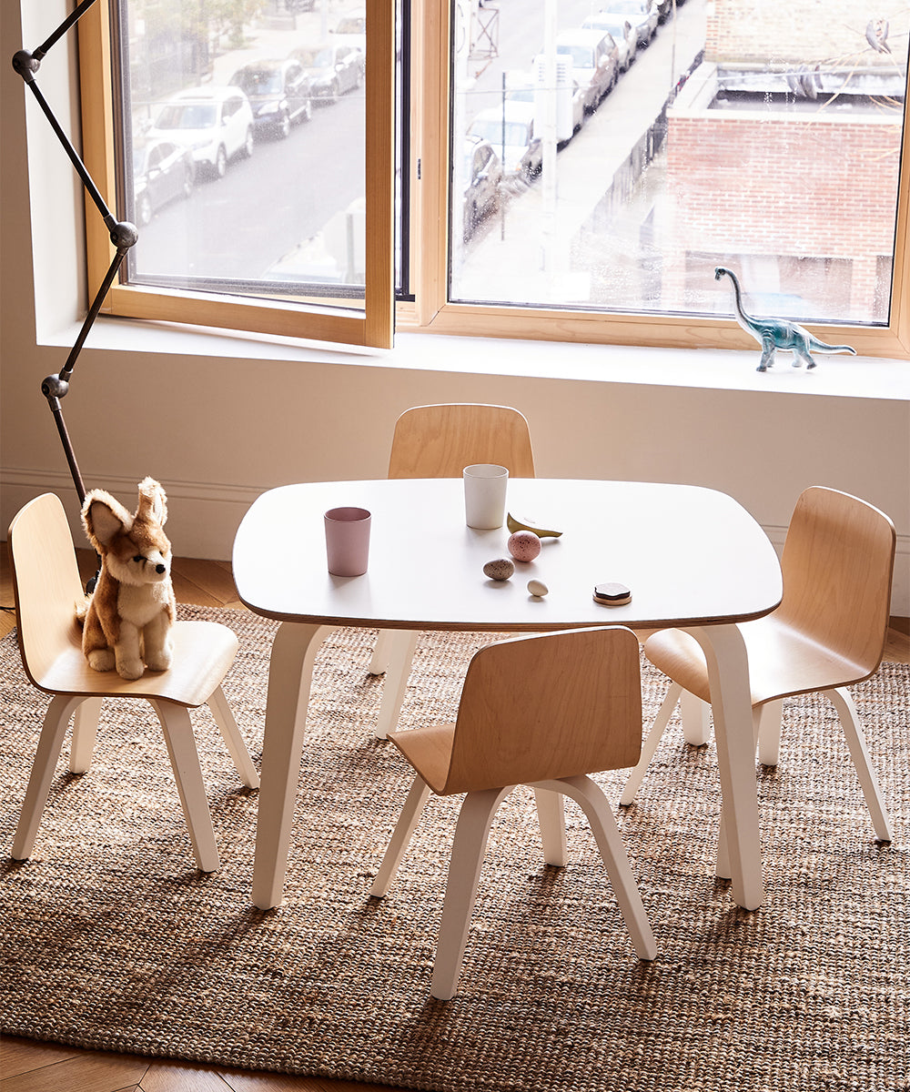 In a sunny room by a large window, theres a childrens table with two sets of Play Chairs, accompanied by a stuffed rabbit, toy dinosaur, cups, and stones.