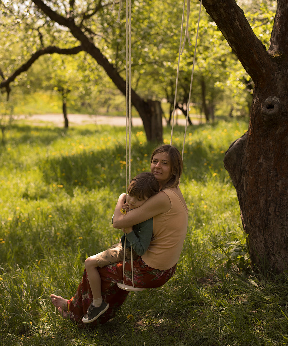 A woman and child sit together on the Le Wooden Swing hanging from a tree in a sunlit field.