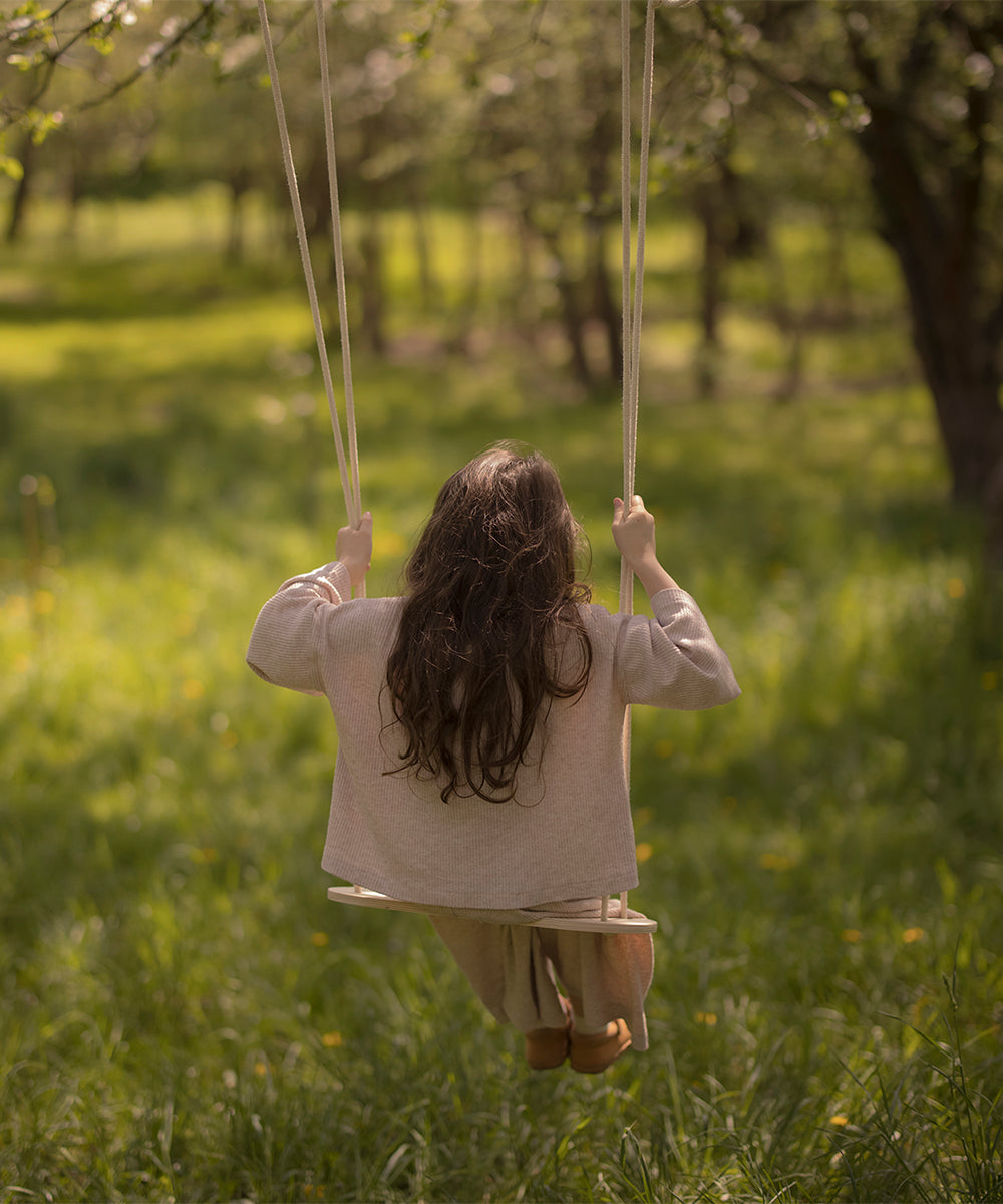 A child with long hair enjoys the Le Wooden Swing, facing away, in a sunlit grassy area surrounded by trees.