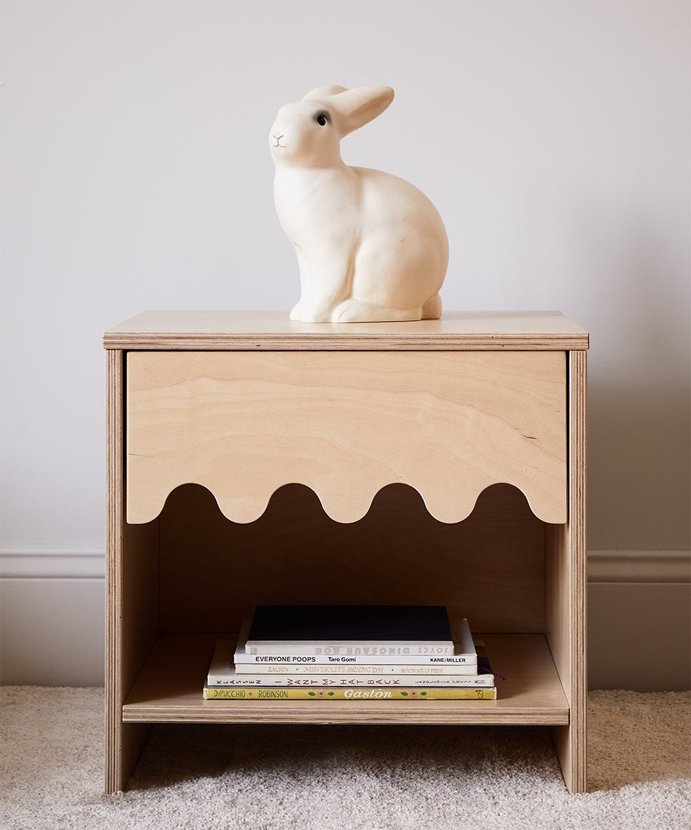 The Moss Nightstand features a scalloped edge and comes topped with a white rabbit figurine, while books are neatly stacked on the lower shelf.