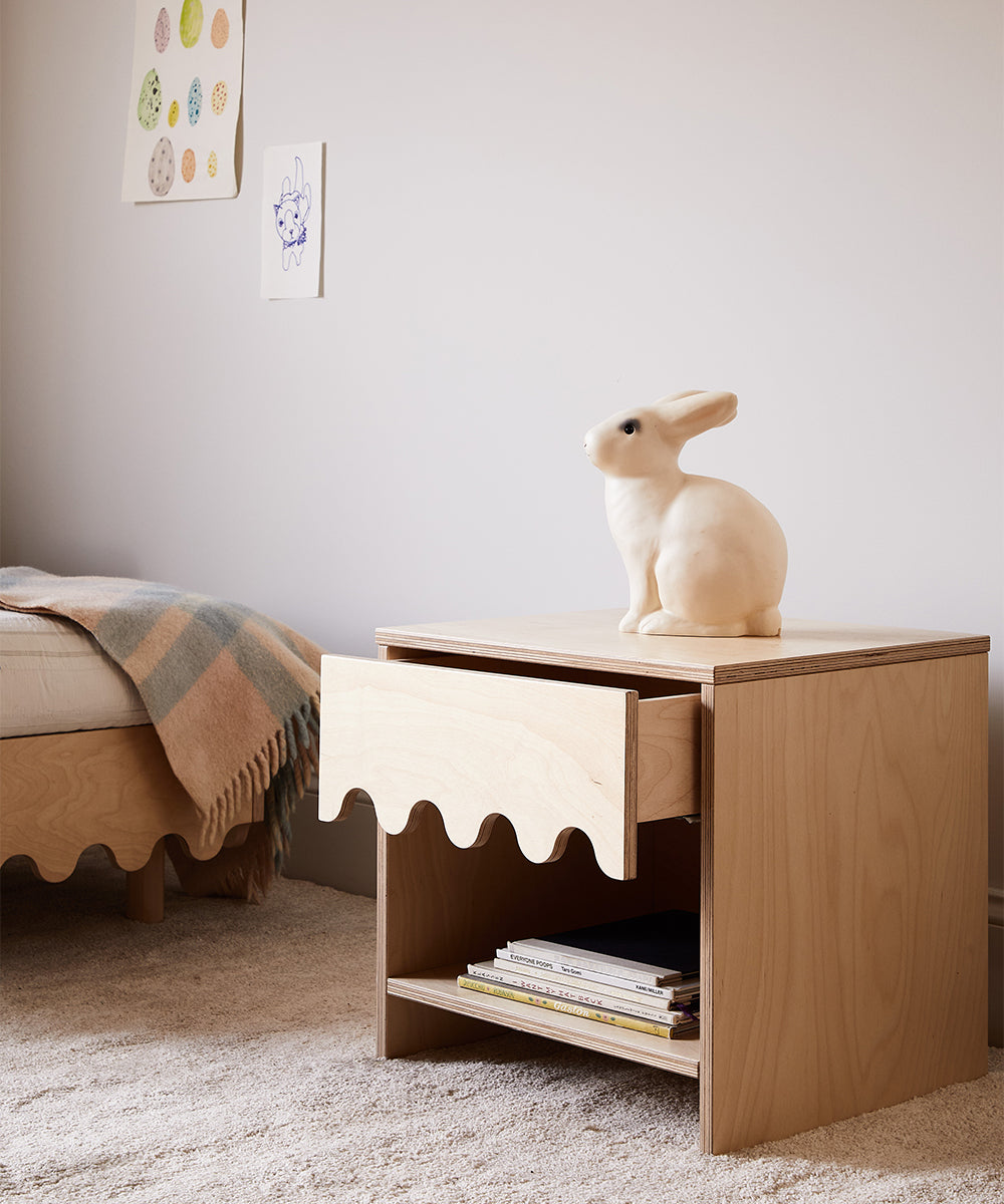 A Moss Nightstand with an open drawer, topped with a rabbit figurine, sits near a bed adorned with a checkered blanket in a cozy room.