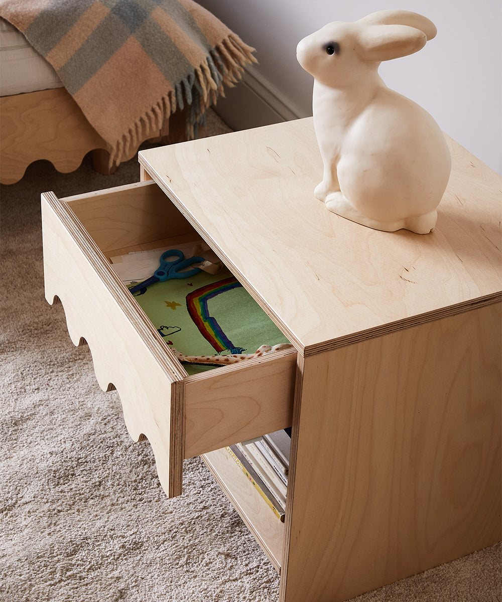 The Moss Nightstand features scalloped edges, an open drawer displaying crafts, and a rabbit figurine on top.