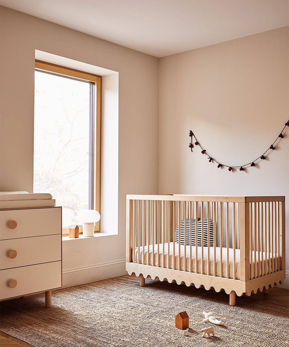 Minimalist nursery featuring the Moss Crib, a dresser, toys on a rug, and a window with a view.