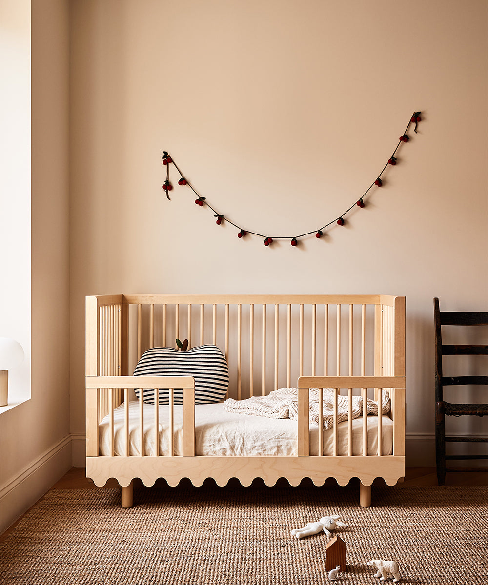 A cozy nursery featuring a Moss Crib Conversion Kit, a striped pillow, wall garland, and a soft brown carpet.