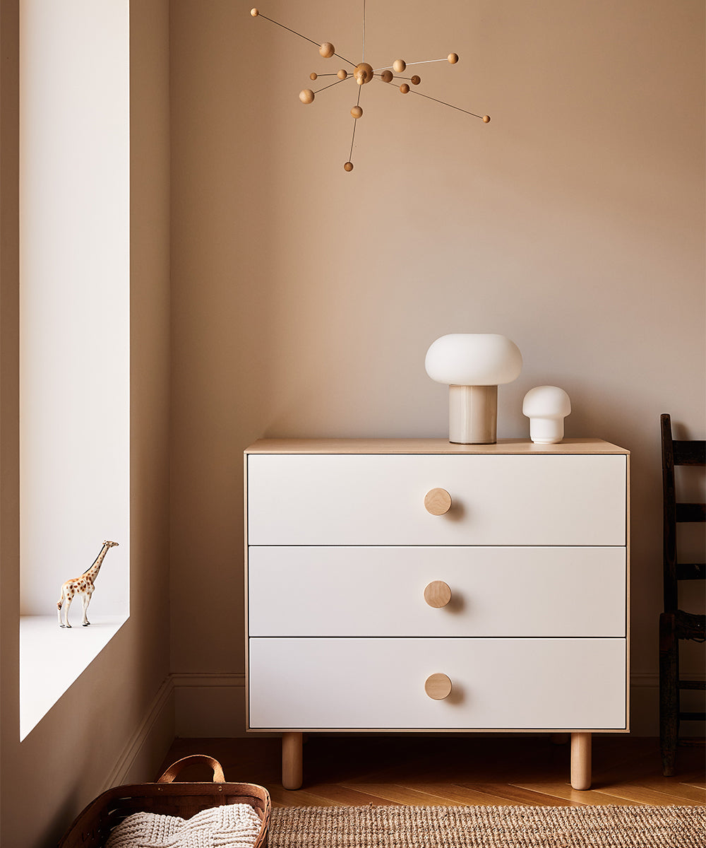 Minimalist room featuring a Dot 3-Drawer Dresser, decorative lamps, wooden mobile, toy giraffe on the windowsill, and wicker basket.