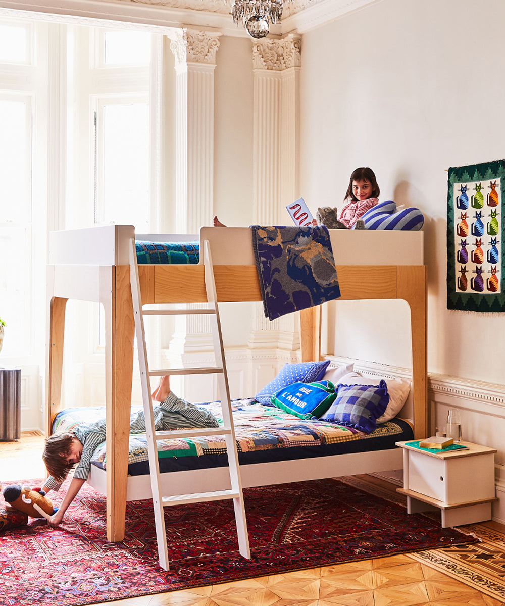 Children playing on a cozy white bunk bed with a colorful rug, featuring a Gingham Ruffle Pillow by the window.