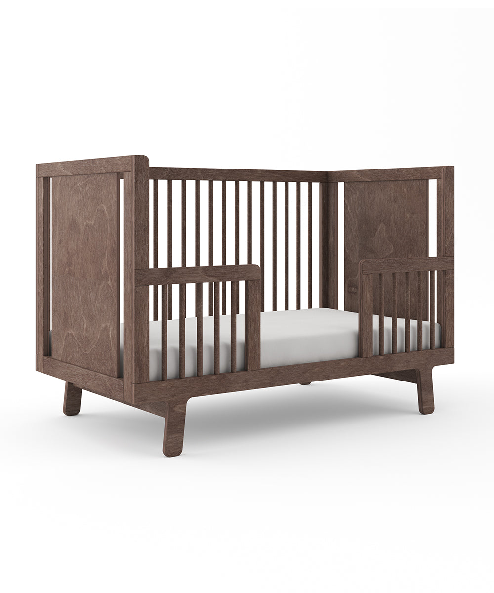 A Sparrow Toddler Bed Conversion Kit featuring a wooden crib with slatted sides and a white mattress, viewed from an angle.