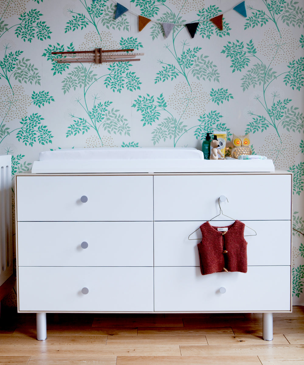 The white dresser, topped with the Convertible XL Changing Station, is decorated with nursery essentials and a sweater on the drawer. Its set against floral wallpaper and features a decorative banner above for a charming touch.