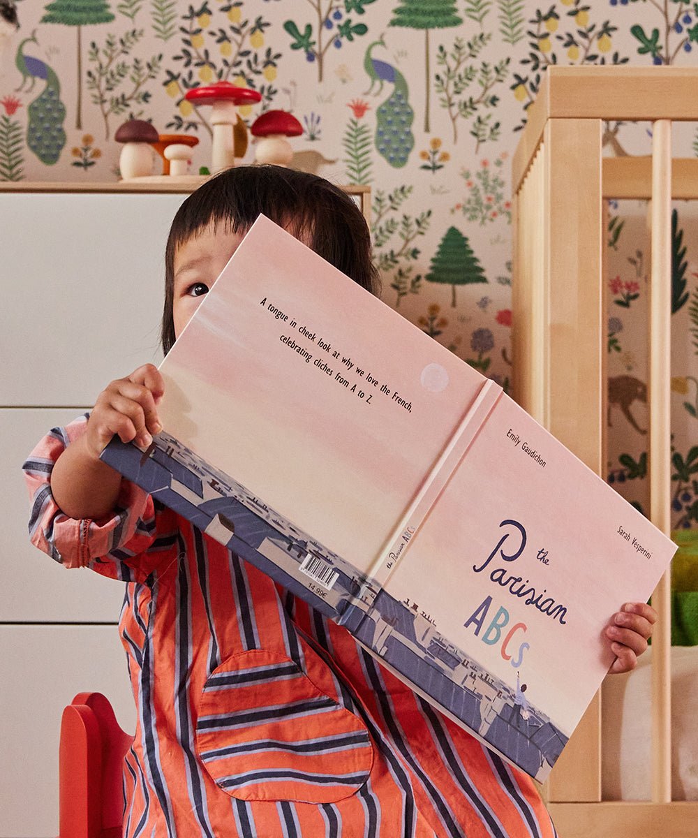 A child sits in a room with patterned wallpaper and a wooden crib, holding a book titled Le Parisian ABCs, partially covering their face.