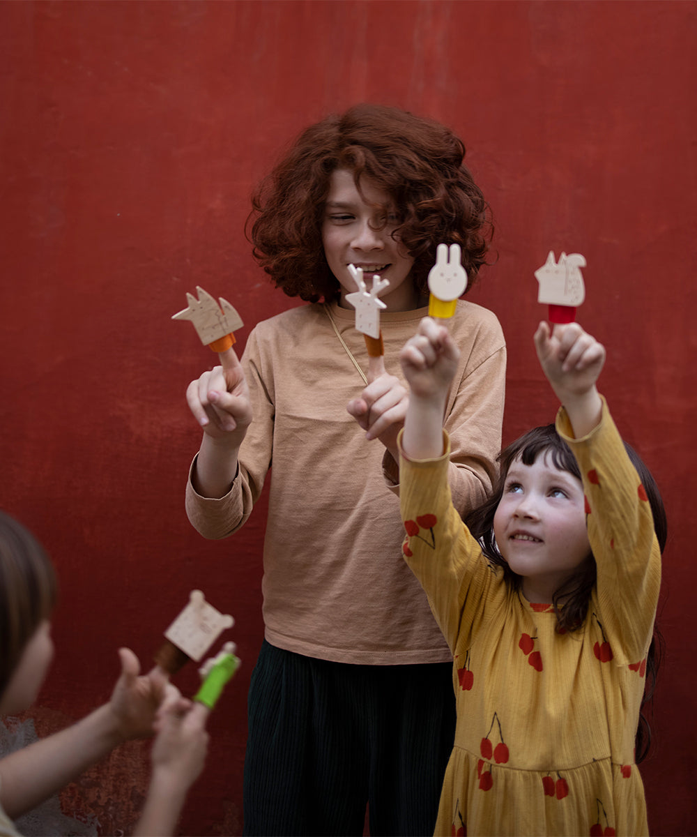 Two children hold Le Bear Finger Puppets against a red background. One child, with curly hair, wears a beige top; the other, with straight hair, sports a yellow dress adorned with cherry prints.