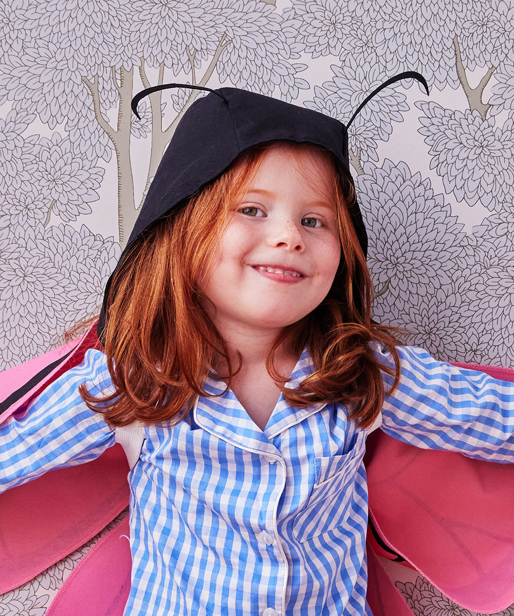 A red-haired child in a checkered shirt, wearing the Le Antennae Hat and butterfly wings, smiles against leafy wallpaper.