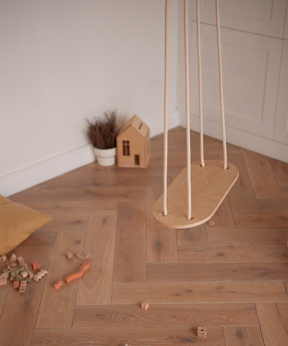 The Le Wooden Swing hangs indoors, surrounded by scattered wooden blocks and a small toy house on a brown herringbone floor.