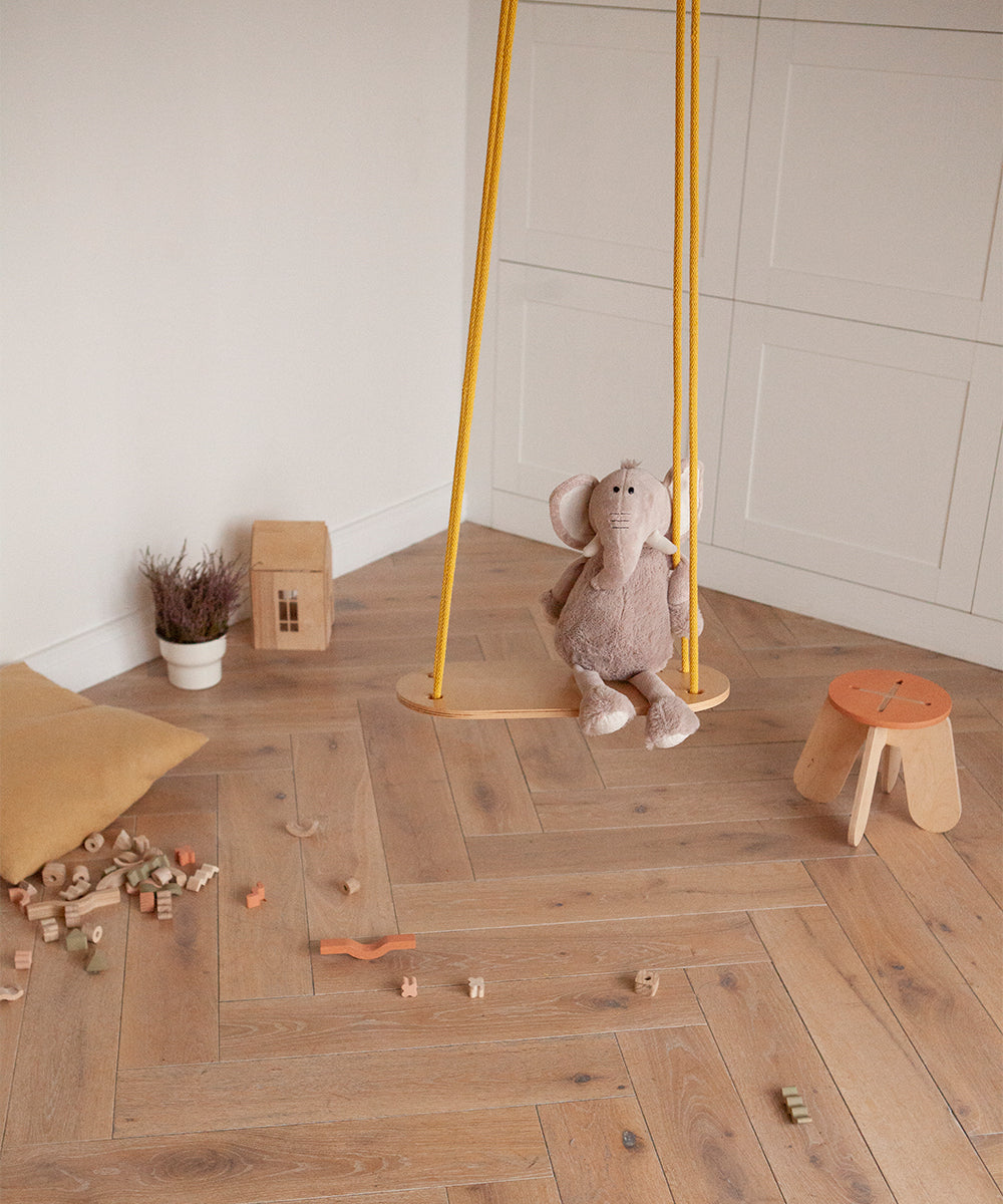 The Le Wooden Swing holds a plush elephant toy indoors. Nearby, a stool, potted plant, wooden house, and scattered wooden blocks rest on the herringbone-patterned wood floor.