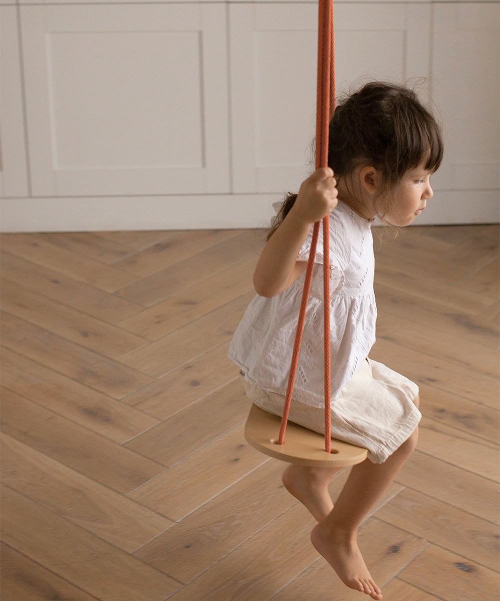 A child sits on a Le Wooden Swing with orange ropes, wearing a white top and beige shorts, in a room with wooden flooring.