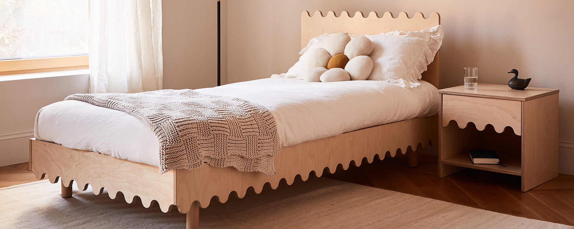 A neatly made bed with a wooden headboard featuring wavy edges, a textured blanket, and decorative pillows. A matching bedside table holds a glass of water and a dark sculpture.