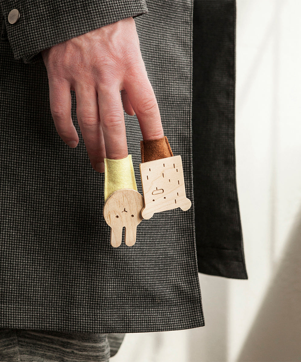 Two fingers adorned with whimsical wooden puppet rings, one is a bunny and the other Le Bear Finger Puppet, both featuring colorful felt details against a gray checkered fabric backdrop.