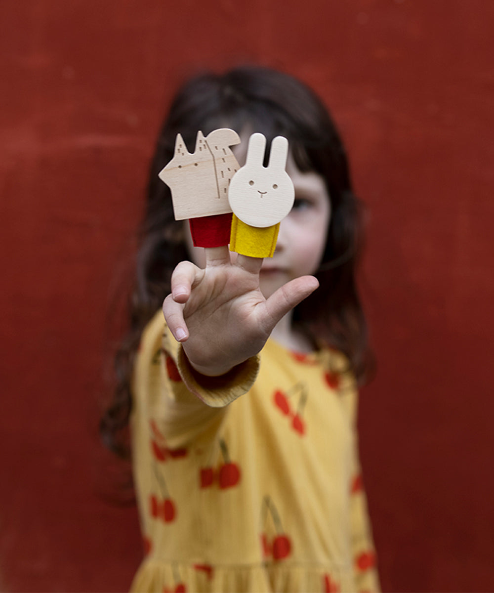 A child holds a wooden Le Squirrel finger puppet in one hand and a rabbit puppet in the other against a red background.