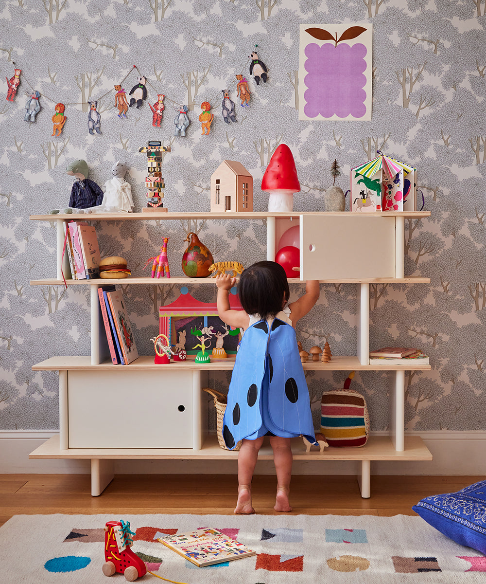 A child wearing Le Blue Butterfly Wings reaches for items on a shelf in a colorful, toy-filled room with patterned wallpaper.