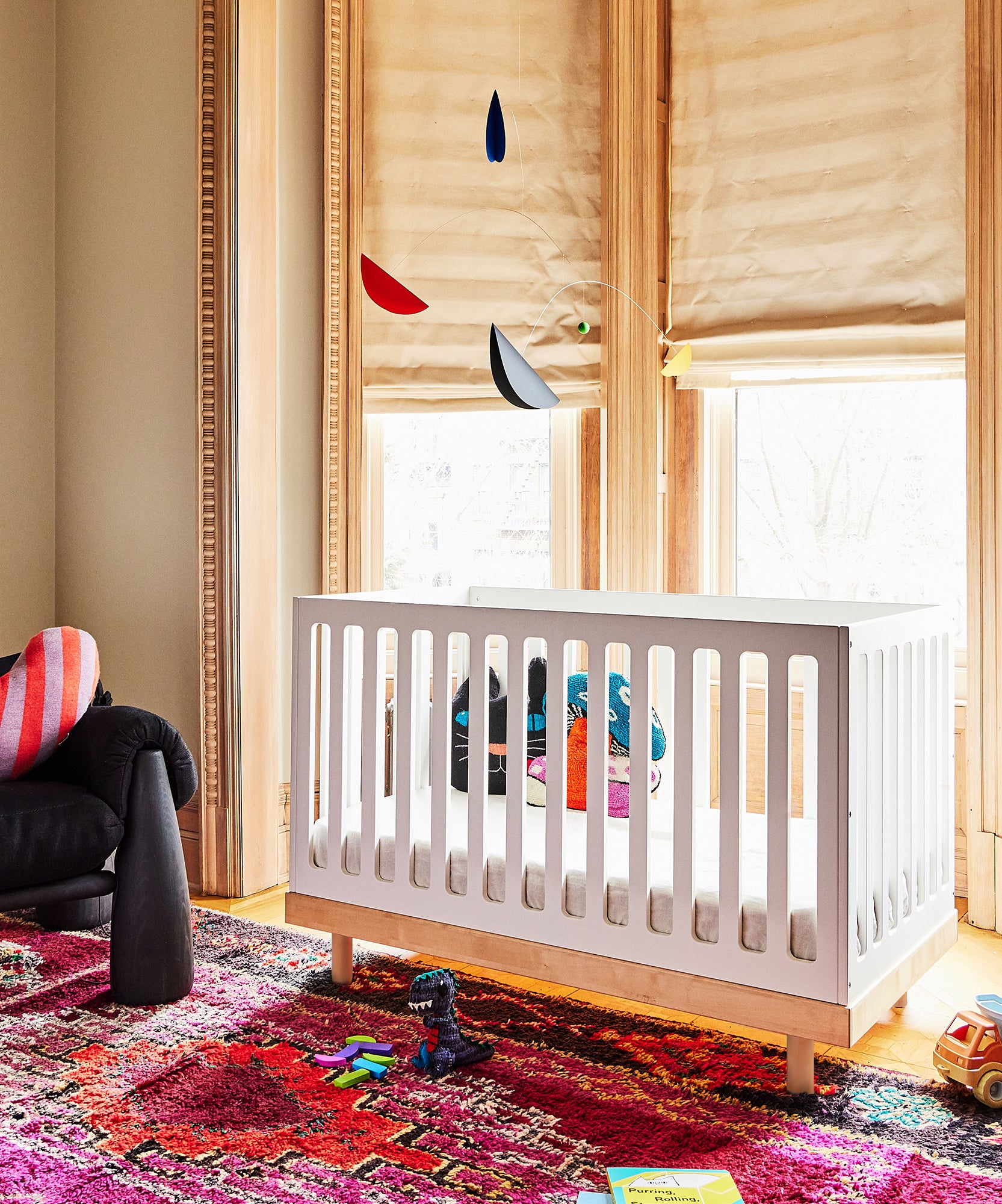 Child's room with a Classic Crib, colorful rug, toys scattered on the floor, and a hanging mobile near large windows.