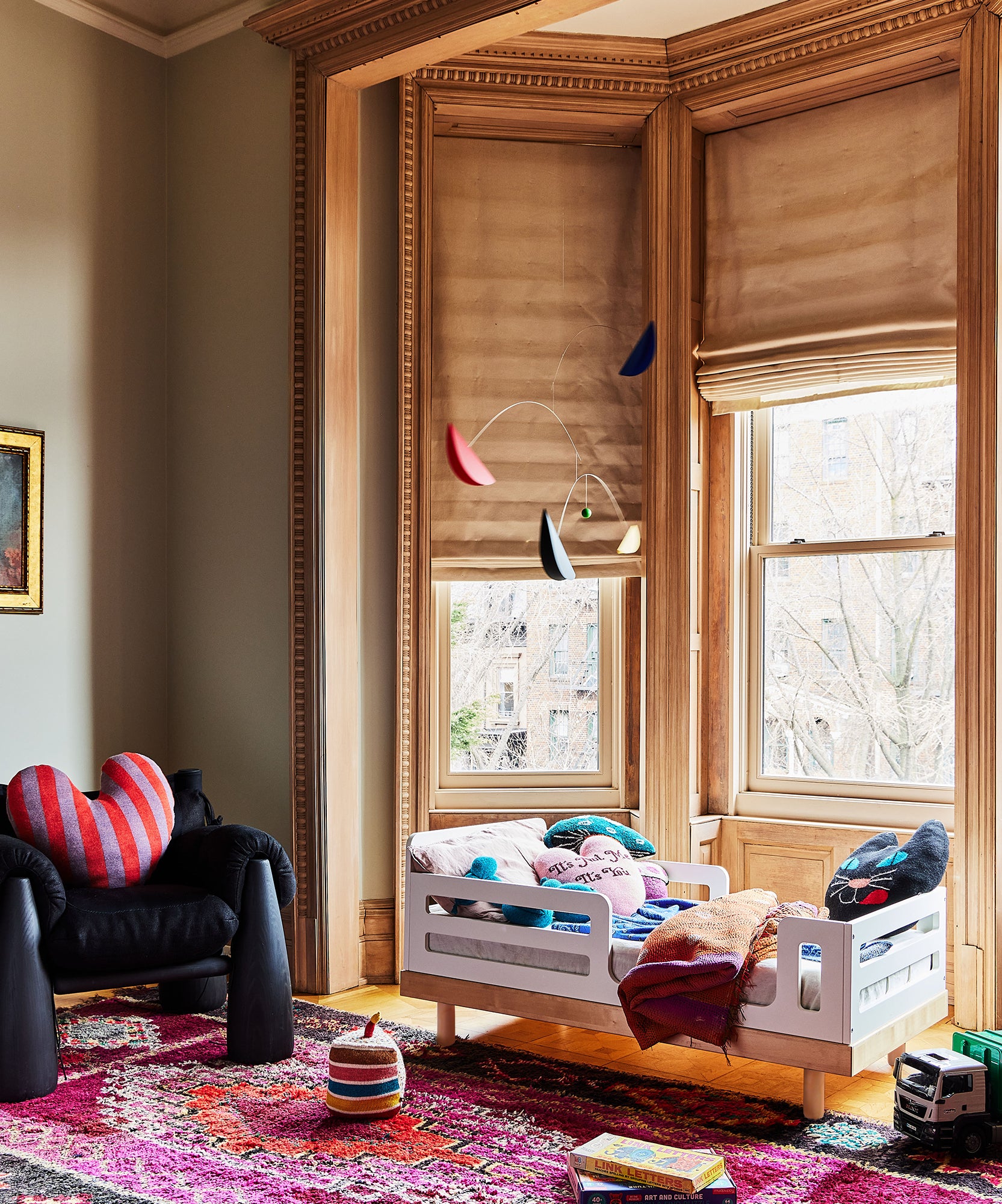 Cozy room with a small Classic/Arbor Toddler Bed, armchair, colorful rug, and large wooden-framed windows with beige blinds.