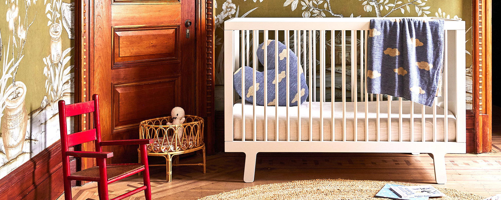 Nursery scene with a white crib, blue pillows, a hanging blue blanket, a small red chair, and a wicker basket with a toy, set on a wooden floor with patterned wallpaper.