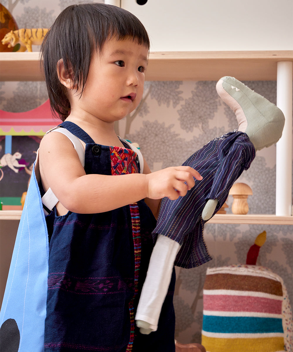 A toddler stands in a playroom holding Le Crocodile, a stuffed animal. The child wears a sleeveless, colorful outfit with toy shelves and wall decor visible behind them.