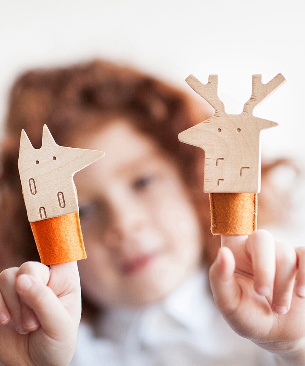 Child holding two wooden finger puppets: Le Deer Finger Puppet and a fox, both with orange felt bases.