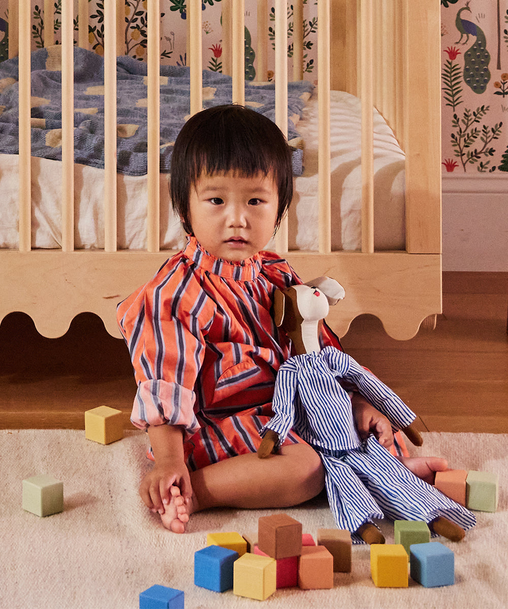 A child in a striped outfit sits on a rug with Le Doggy, surrounded by colorful blocks, in front of a wooden crib.