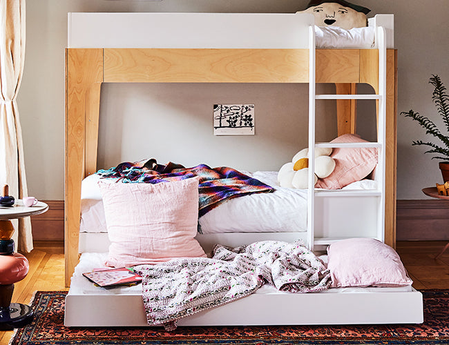 A cozy room with a wooden bunk bed, pink pillows, a colorful blanket, and a small ladder. A plant is visible in the corner, and a rug covers the wooden floor.