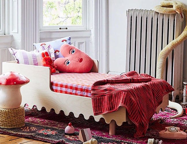 Childs bedroom with a wooden bed, checkered bedding, and red blanket. Plush heart-shaped pillow, toys, and a radiator are visible. Decor includes a faux mushroom and a colorful rug.