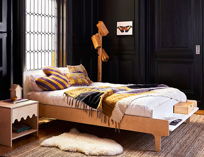 Bedroom with a wooden bed and side table. The bed has striped pillows and a multi-colored blanket. A sheepskin rug lies on the floor. A tree-shaped lamp and butterfly art adorn the room.