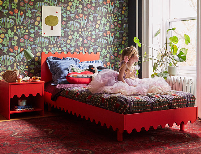 Child in a pink dress sits on a red bed with a quilt, against floral wallpaper. A plant is by a large window. The room has a red nightstand and a red patterned rug.