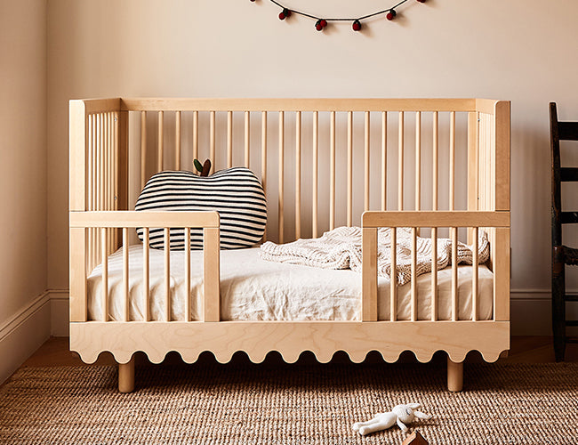 Wooden crib with scalloped bottom edge, striped apple-shaped pillow, and a blanket inside. A rug is on the floor, with a stuffed toy on it. A decorative garland hangs on the wall.