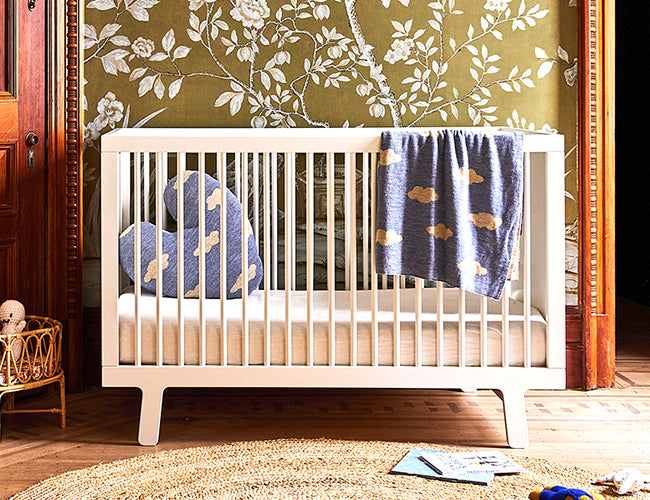 White crib with a heart-shaped blue pillow and a blue blanket draped over the side with cloud patterns, set against a floral wallpaper background.