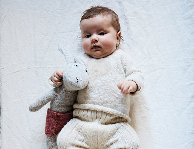 Baby lying on a white blanket, holding a gray stuffed animal with pink pants. The baby is wearing a cream-colored knitted outfit.