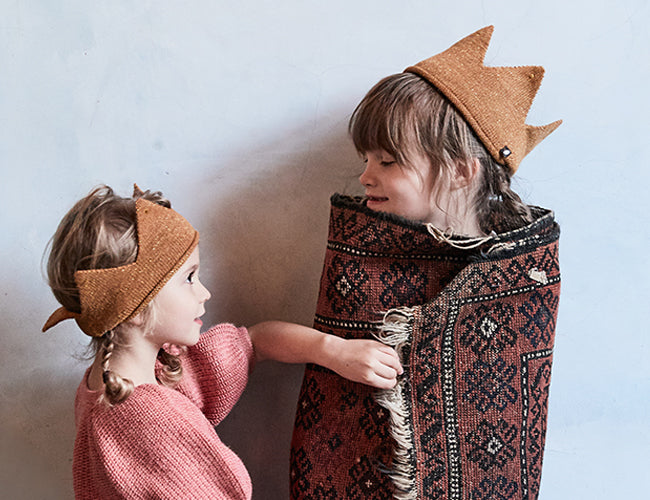 Two children wearing golden crowns and wrapped in a patterned blanket stand against a neutral background.