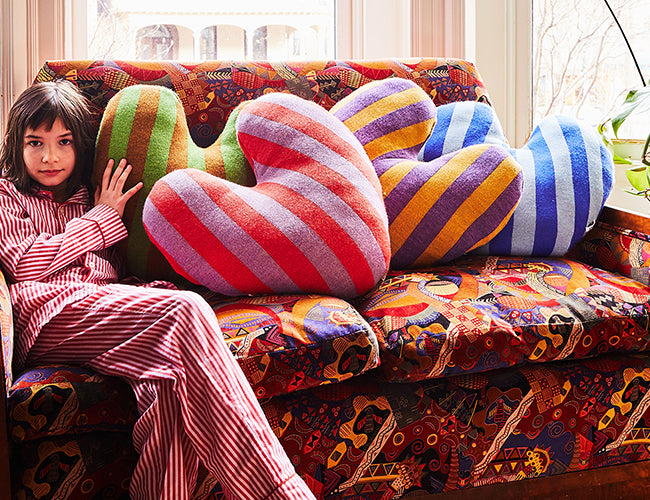 A person in striped pajamas sits on a colorful patterned couch with four large, striped heart-shaped pillows in various colors.