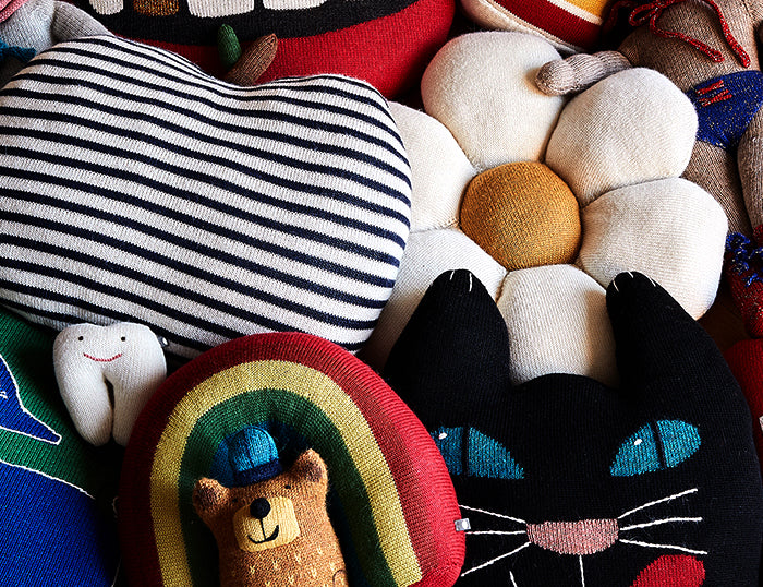 A collection of colorful knitted pillows, including a striped heart, a daisy, a tooth, a rainbow with a bear, and a black cat face.