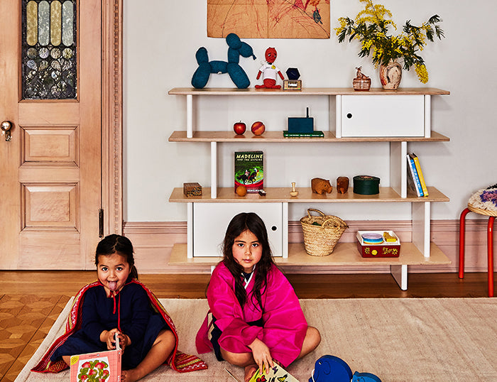 Two children sitting on a rug with books, in a room with shelves displaying toys and decor.