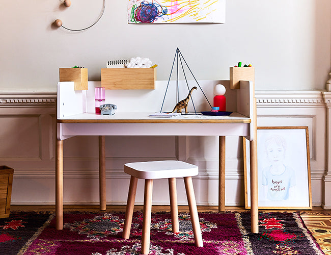 Childs desk with assorted toys and art supplies, a small stool, framed artwork on the floor, and a colorful rug in a room with wainscoting.