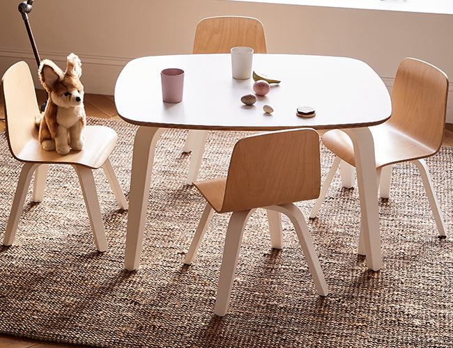 Childrens playroom with a square table, four wooden chairs, and a plush dog toy on one chair, on a woven rug. Various small toys and cups are on the table.