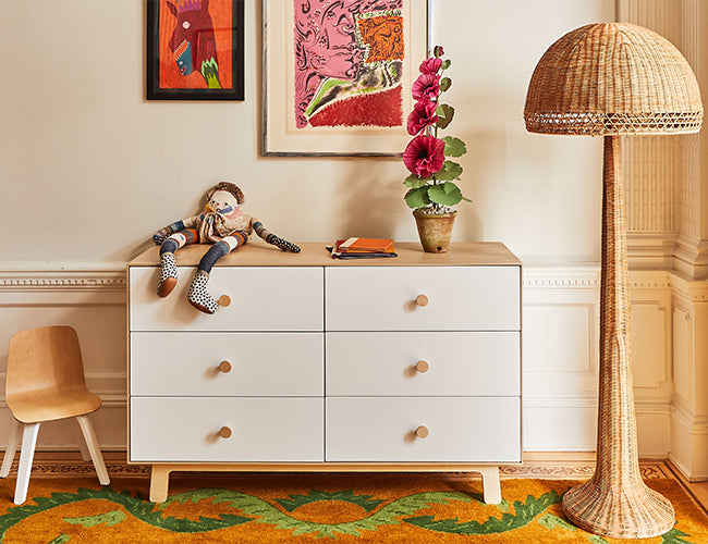 A white dresser with six drawers, a doll seated on top with an orange book, alongside a wicker floor lamp, a small wooden chair, and framed art hanging above.