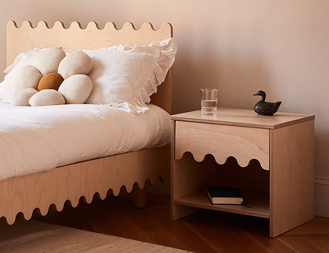 Wooden bed with a white duvet set and decorative pillows. Next to it, a matching wooden nightstand holds a small black duck figurine, a glass of water, and a closed book.