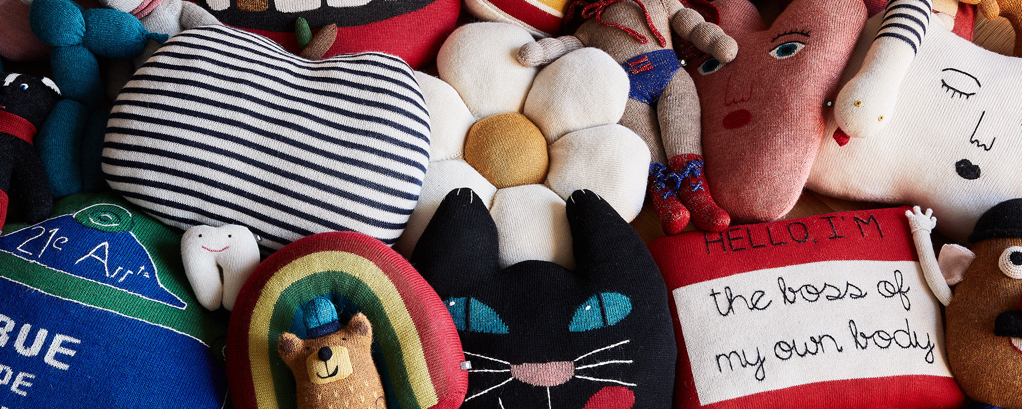 A collection of colorful, whimsical stuffed toys including a striped apple, a cat, a rainbow, and others with various shapes and designs.