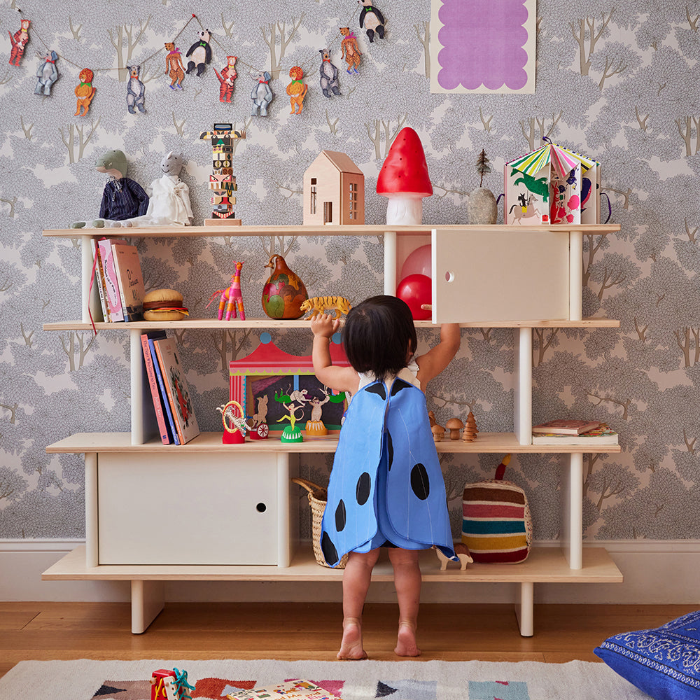 A toddler in a blue dress with black dots reaches up to a white shelf filled with toys and books, in a room with patterned wallpaper and playful decorations.