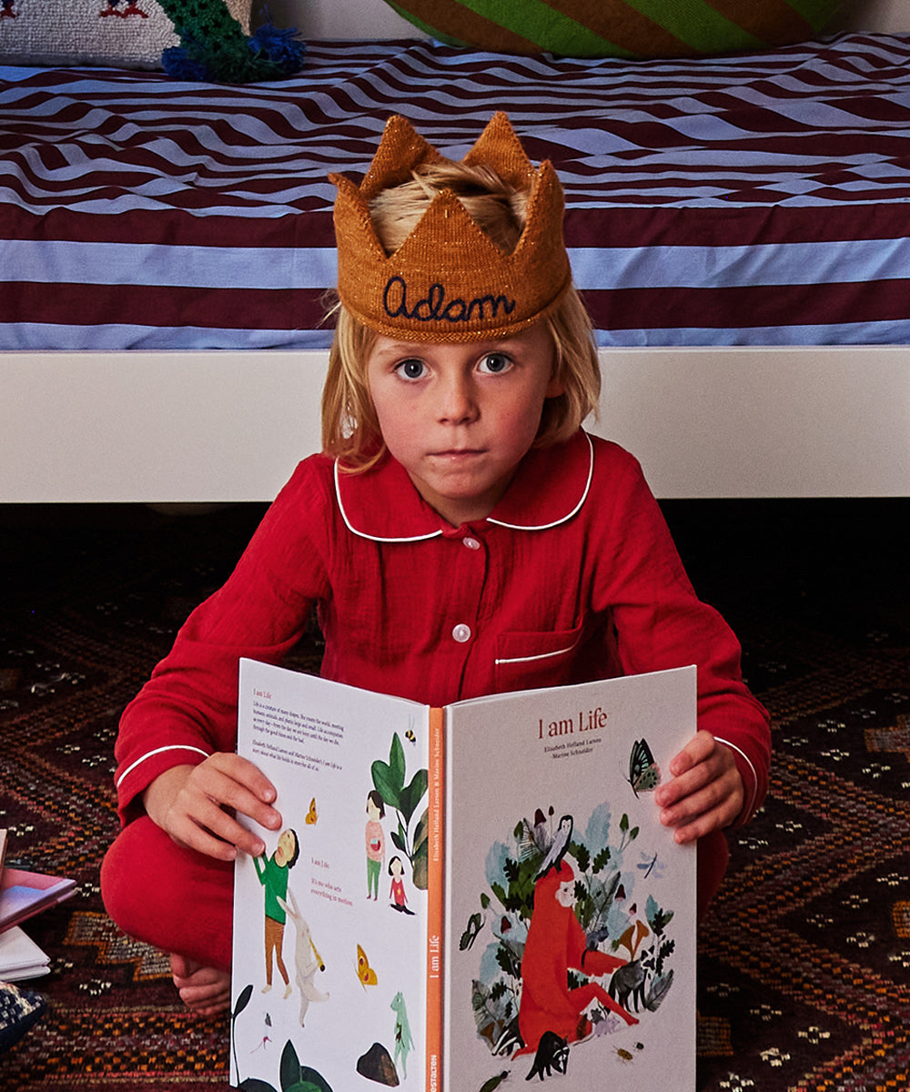 A child in red pajamas, wearing a crown labeled Adam, sits on the floor with an open copy of Le Book - I Am Life in front of him, near a bed.