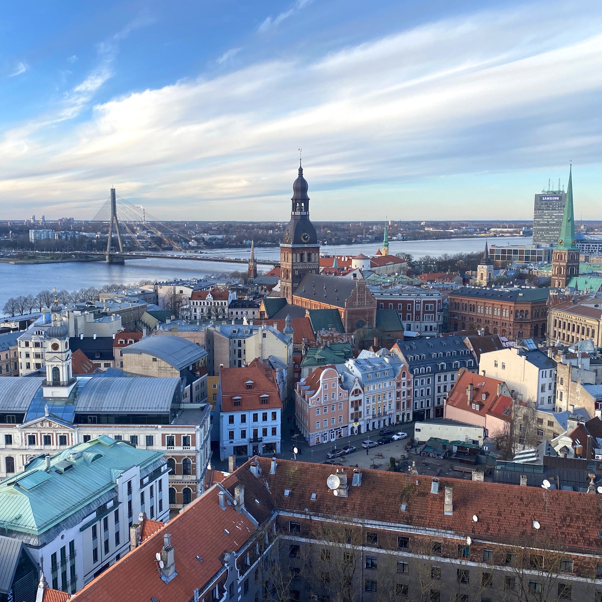 Aerial view of a historic European city with a river in the background, featuring a prominent church tower and a mix of colorful buildings. A bridge spans the river in the distance under a cloudy sky.