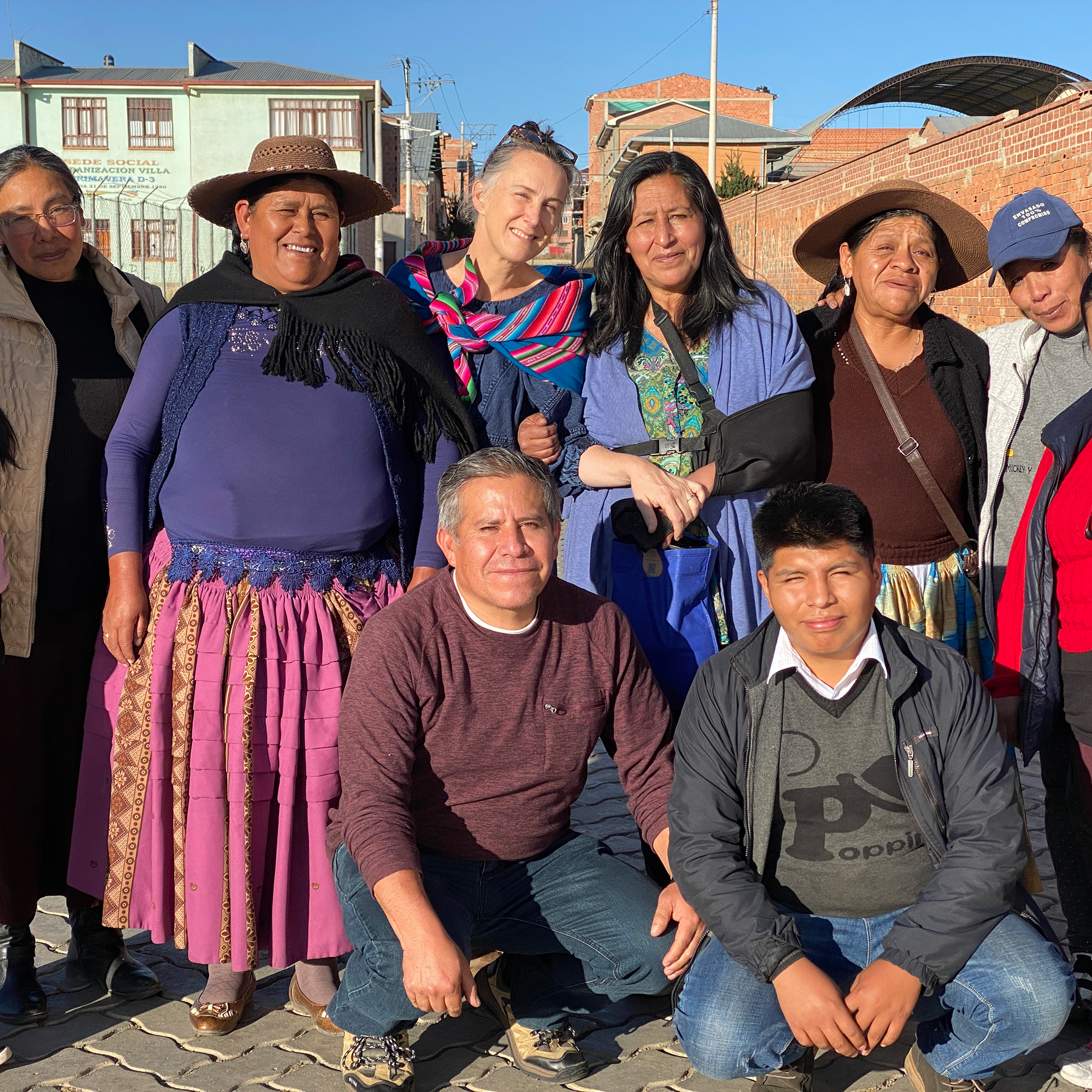 A group of people, including both men and women, pose together outdoors on a sunny day. Some are wearing traditional clothing and hats. Buildings and a blue sky are in the background.