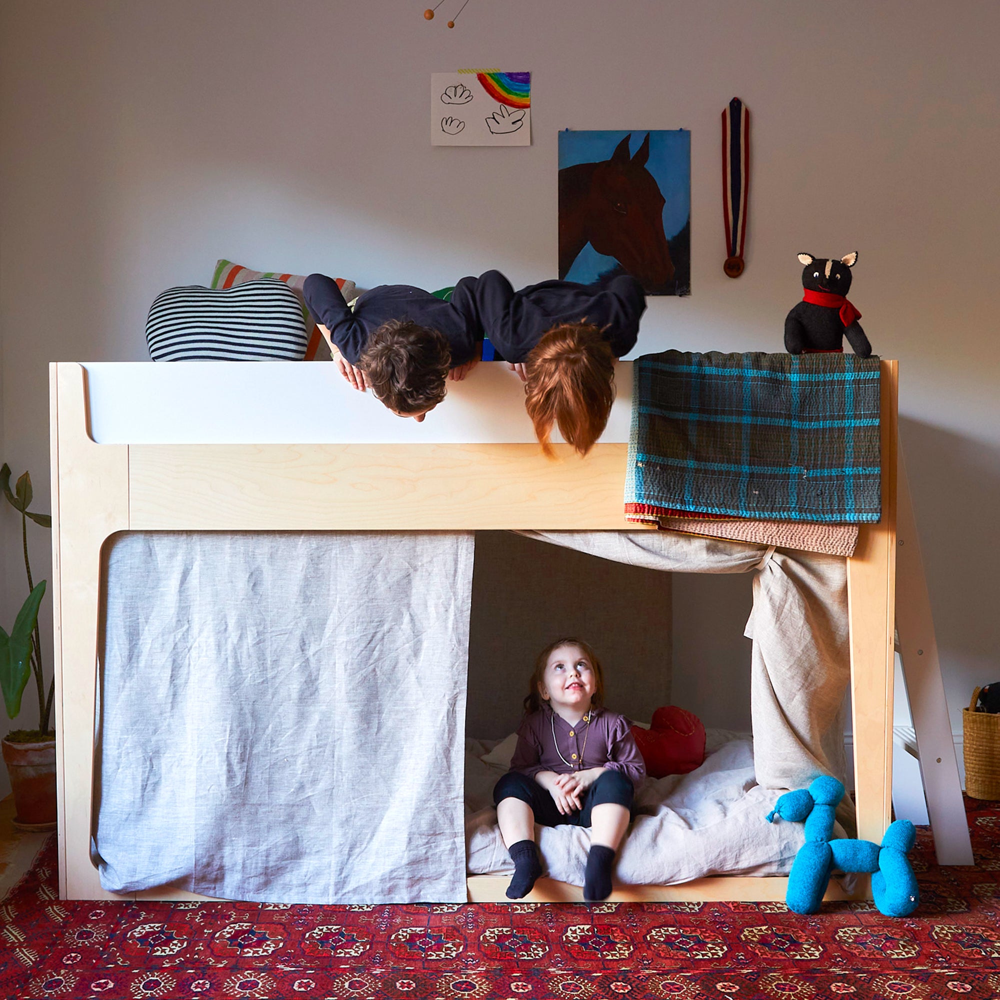 Two children lie on the top bunk, looking down at a child sitting on the lower bunk bed. A plush toy dog and decorations are in the room.